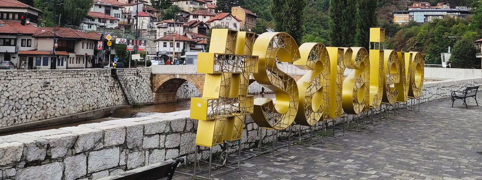 Eine große goldene 3D-Installation mit der Aufschrift „#Sarajevo“ steht auf einer Promenade entlang eines Flusses. Im Hintergrund sind traditionelle Gebäude mit roten Dächern und eine Steinbrücke über den Fluss zu sehen. Umgeben von Hügeln und Bäumen vermittelt die Szene ein Gefühl von historischer und moderner Kultur.