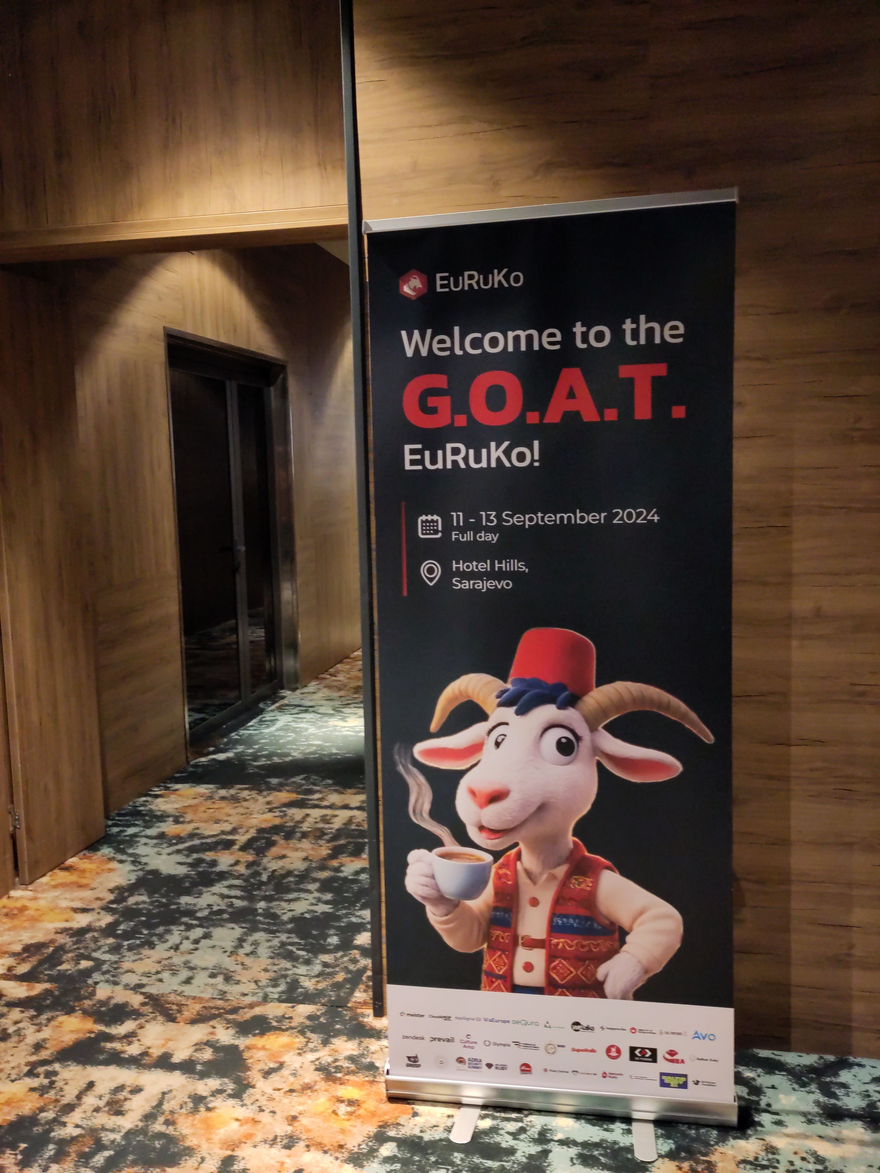 Ein Roll-Up-Banner mit dem Logo von „EuRuKo“ und dem Text „Welcome to the G.O.A.T. EuRuKo!“ zeigt eine illustriere Ziege, die eine Kaffeetasse hält und traditionelle Kleidung trägt. Der Banner kündigt eine Konferenz an, die vom 11. bis 13. September 2024 in Sarajevo stattfindet.